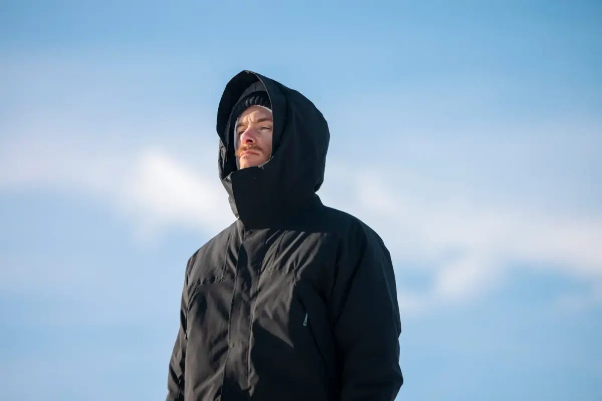 Mann in schwarzem Parka mit aufgesetzter Kapuze vor blauem Winterhimmel