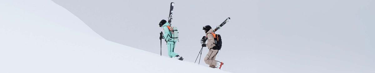 Zwei Personen in Skibekleidung steigen mit geschulterten Ski einen verschneiten Hang hinauf