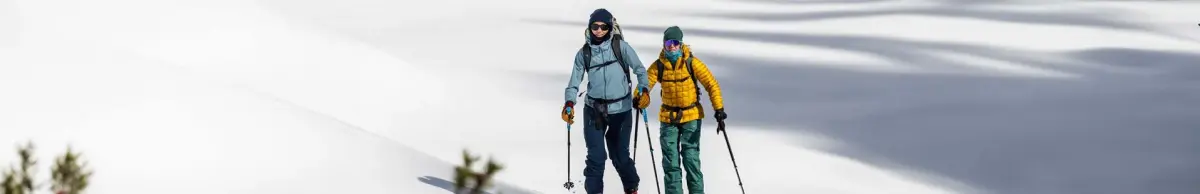 Zwei Skitourengeher in blauer und gelber Jacke steigen einen sonnigen Schneehang hinauf