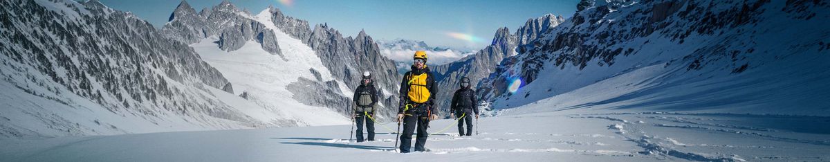 Drei Bergsteiger am Seil gehen über einen verschneiten Gletscher, dahinter felsige, schneebedeckte Gipfel