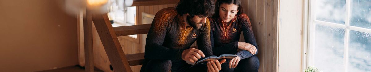 Ein Mann und eine Frau in dunklen Funktionsshirts mit orangefarbenem Muster sitzen auf einer Holztreppe und lesen gemeinsam in einem Heft