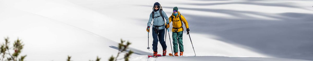 Zwei Personen auf Skitour steigen mit Stöcken durch eine verschneite Mulde, eine in hellblauer Jacke, eine in gelber Steppjacke