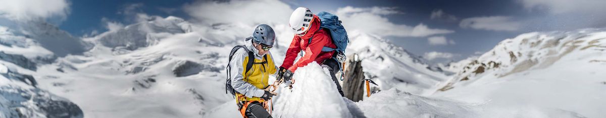Zwei Bergsteiger mit Helm an einem verschneiten Grat, einer in gelb-grauer Jacke mit Seil in der Hand, einer in Rot mit blauem Rucksack, dahinter Gletscher