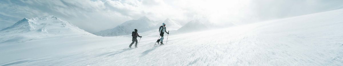 Zwei Skitourengeher steigen mit Rucksack und Stöcken über einen verschneiten Hang, dahinter Berggipfel im Nebel