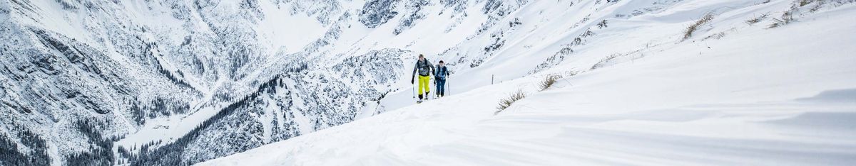 Zwei Skitourengeher steigen über einen verschneiten Grat auf, vorn einer in gelber Hose, dahinter tief verschneite Berghänge