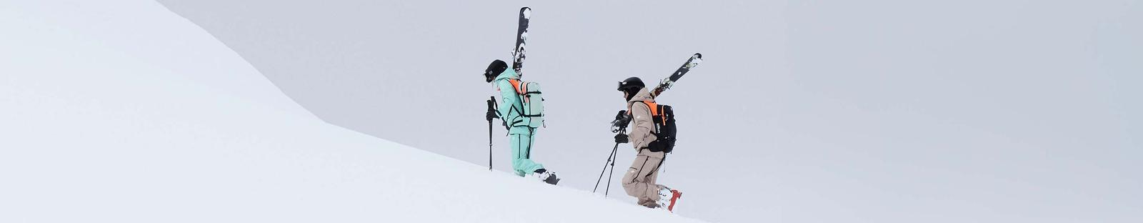Zwei Personen in Skibekleidung steigen mit geschulterten Ski einen verschneiten Hang hinauf