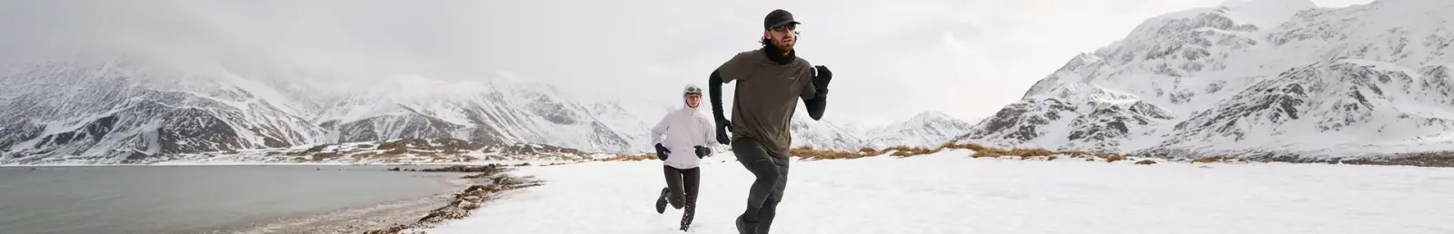 Zwei Läufer auf Schnee an einem Bergsee vor verschneiten Gipfeln