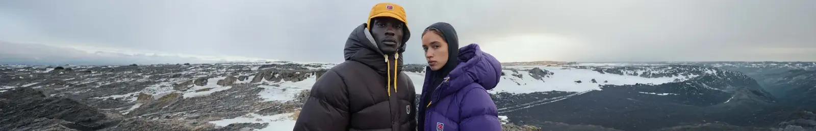 Zwei Personen in Fjällräven-Daunenjacken, braun und lila, vor einer schneebedeckten Vulkanlandschaft