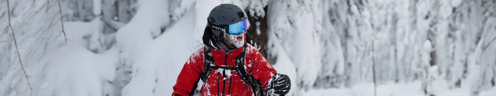 Skifahrer in roter Jacke mit schwarzem Smith-Helm und blau verspiegelter Skibrille, Bart und Kragen voller Schnee, im tief verschneiten Wald