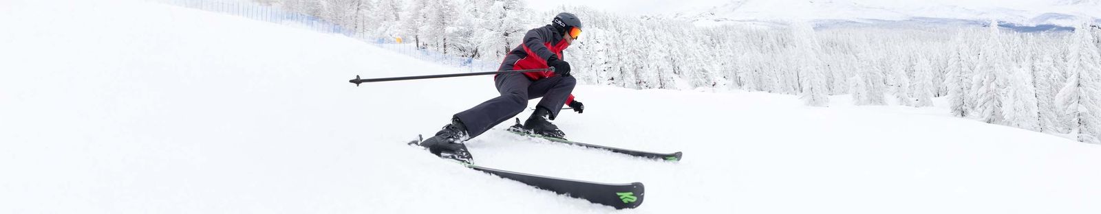 Skifahrer in rot-schwarzer Jacke legt sich auf frisch verschneiter Piste in die Kurve, dahinter verschneiter Wald