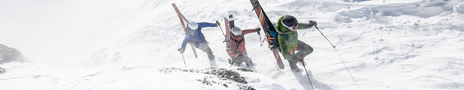 Drei Personen mit Helmen steigen mit Ski und Snowboard am Rucksack einen verschneiten, windigen Grat hinauf