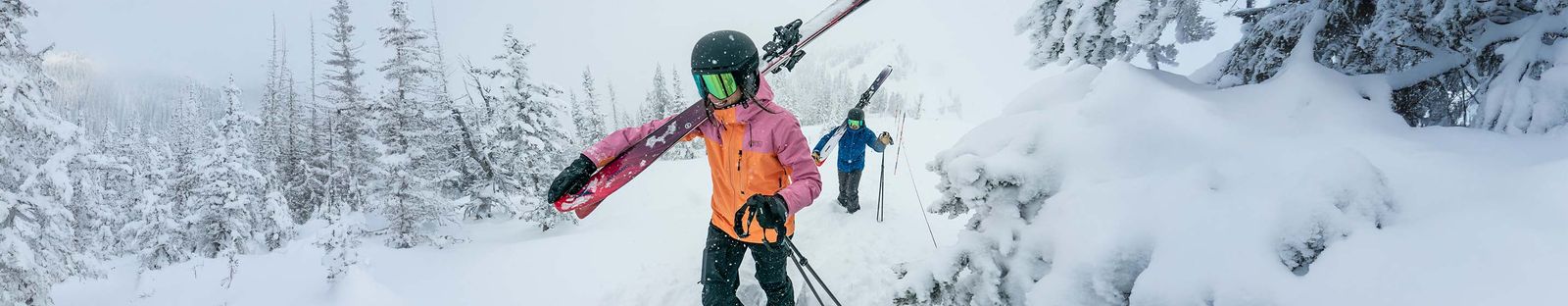Skifahrerin mit schwarzem Helm, verspiegelter Skibrille und orange-rosa Jacke trägt ihre Ski auf der Schulter durch tief verschneiten Wald, hinter ihr ein Skifahrer in Blau
