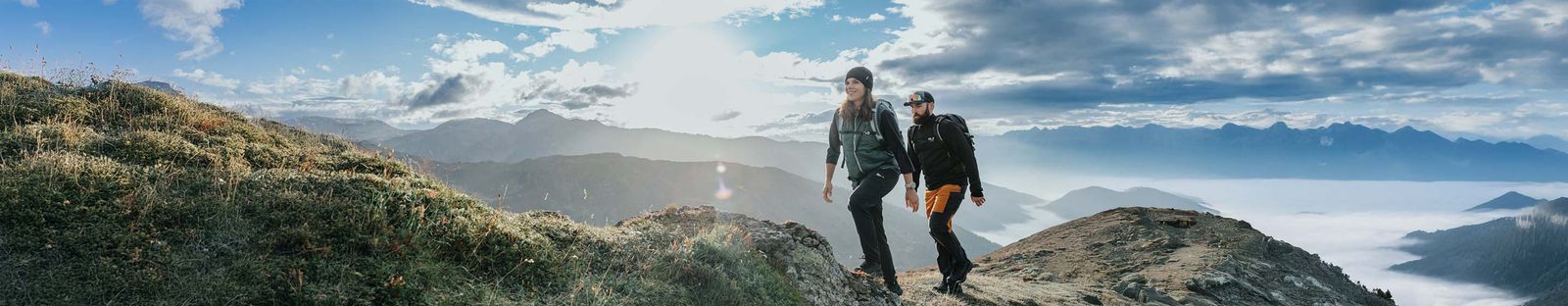 Zwei Wandernde mit Rucksäcken und Mützen auf einem grasigen Bergrücken, im Tal liegt Nebel, dahinter Bergketten im Gegenlicht