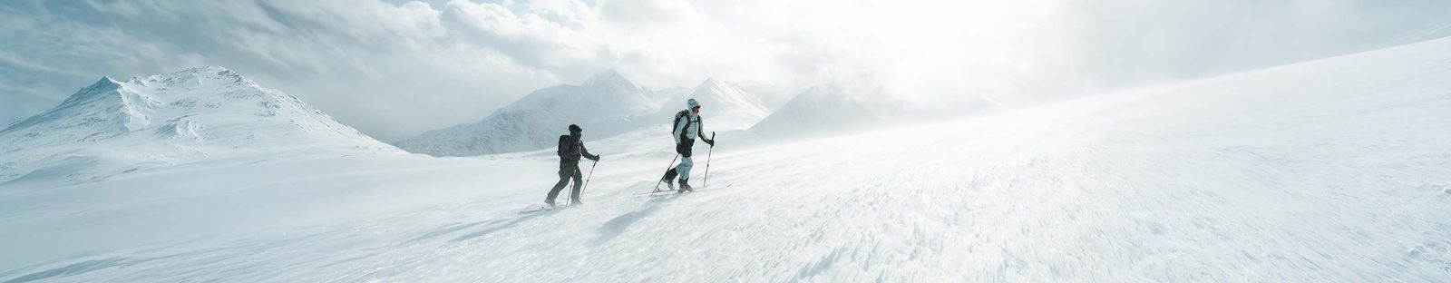 Zwei Skitourengeher steigen mit Rucksack und Stöcken über einen verschneiten Hang, dahinter Berggipfel im Nebel