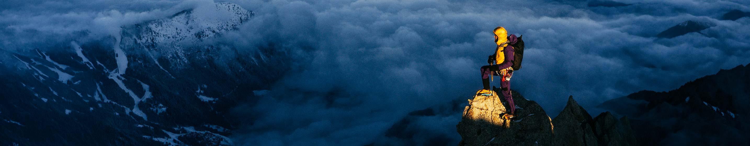 Bergsteiger in gelber Jacke von Montura steht im Abendlicht auf einem Felsgipfel über dem Wolkenmeer