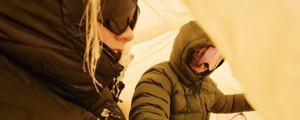 Zwei Personen in Haglöfs Daunenjacken mit Kapuze und Skibrille im gelben Zelt