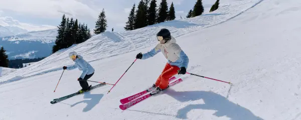 Zwei Skifahrerinnen in Helly-Hansen-Skijacken fahren nebeneinander eine Piste hinunter
