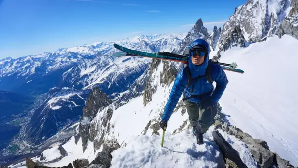 Bergsteiger in blauer Mountain Equipment Xenith Daunenjacke mit Ski auf dem Rucksack steigt über einen verschneiten Felsgrat