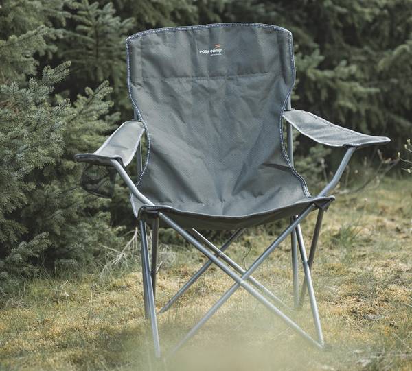 Grauer Klappsessel mit Armlehnen und Getränkehalter steht im hohen Gras vor Nadelbäumen