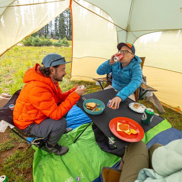 Zwei Personen in Daunenjacken sitzen unter einem Tarp auf niedrigen Stühlen an einem flachen Campingtisch und essen