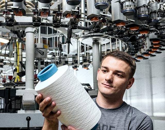 Ein Mann im grauen T-Shirt hält eine große weiße Garnspule vor einer Rundstrickmaschine