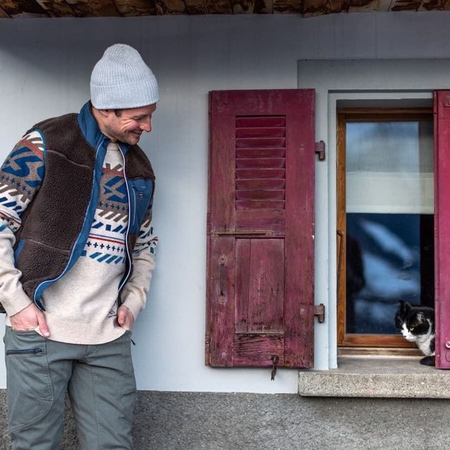 Mann in brauner Fleeceweste über gemustertem Pullover und grauer Mütze vor einem Haus mit roten Fensterläden