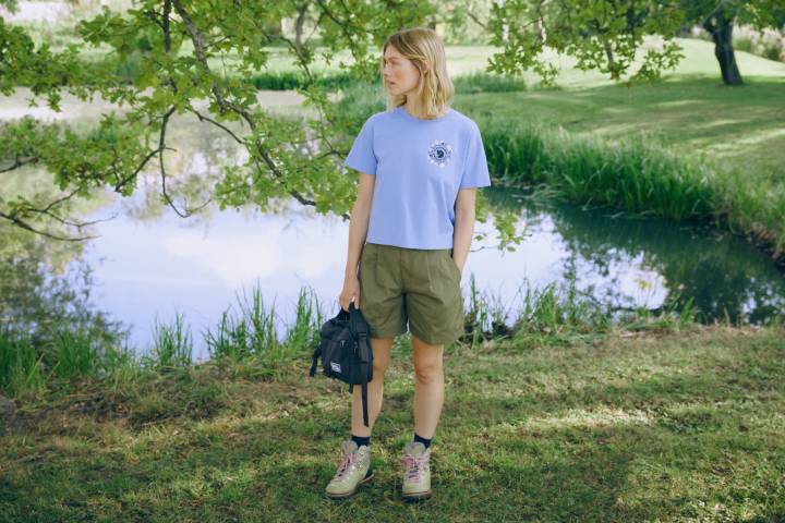 Frau in hellblauem T-Shirt mit Aufdruck und olivfarbenen Shorts steht mit einer Tasche in der Hand an einem Teich unter Bäumen
