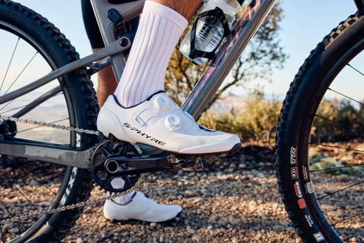 Fuß in weißer Socke und weißem Radschuh mit Drehverschlüssen auf dem Klickpedal eines Mountainbikes, darunter Schotter