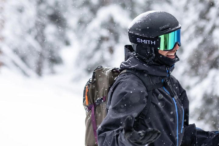 Lachende Person mit schwarzem Smith-Helm und verspiegelter Skibrille, dunkler Jacke und Rucksack im Schneefall vor verschneiten Bäumen