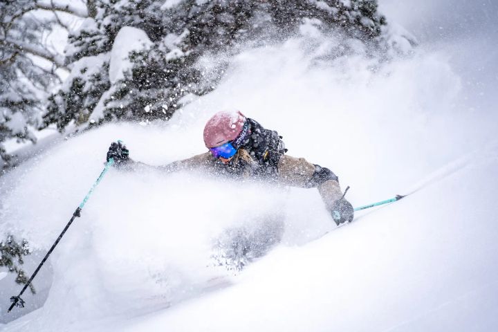 Skifahrer mit rosafarbenem Helm und blau verspiegelter Skibrille fährt durch tiefen Pulverschnee, um ihn herum staubt der Schnee auf