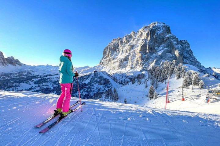 Skifahrerin in pinker Skihose und türkiser Jacke auf einer präparierten Piste, dahinter ein verschneiter Felsberg in den Dolomiten