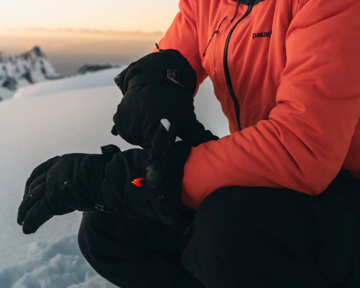 Person in roter Jacke kniet im Schnee und zieht schwarze beheizbare Handschuhe an, ein rotes Licht zeigt die Heizung an