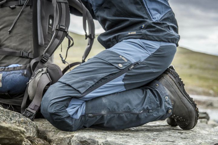 Person in blau-grauer Trekkinghose aus G-1000 kniet auf einem Felsen, neben ihr ein Trekkingrucksack