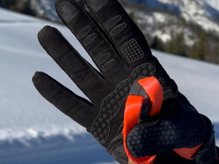 Hand in schwarzem Handschuh mit Noppen auf der Innenseite zieht an einer orangefarbenen Lasche, im Hintergrund Schnee