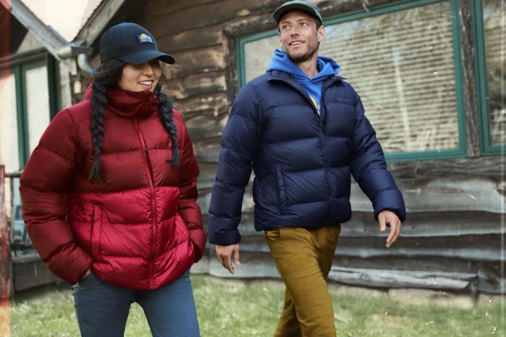 Frau in roter und Mann in dunkelblauer Steppjacke gehen vor einer Holzhütte über eine Wiese