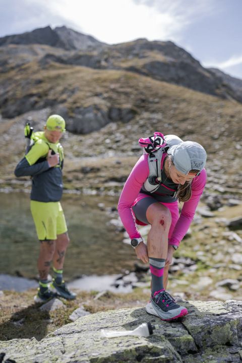 Trailläuferin in Pink zieht am Ufer eines Bergsees ihre Socke hoch, daneben ein Läufer in gelber Hose, beide mit Laufwesten auf Felsen