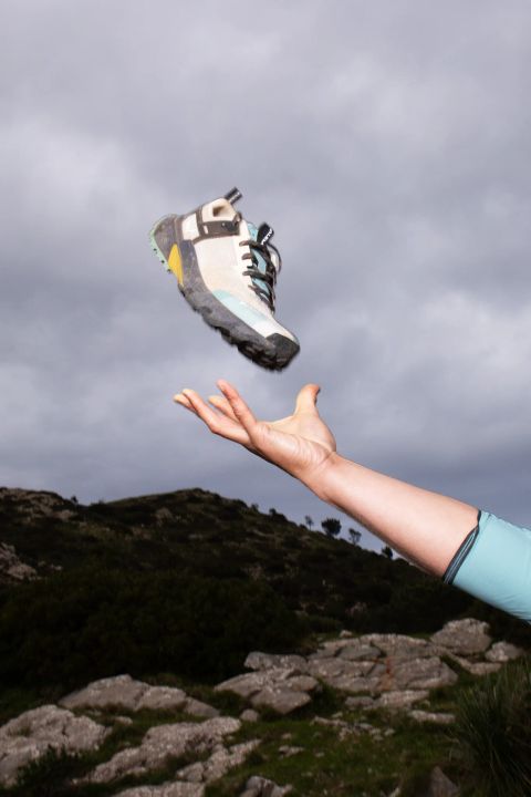 Hand wirft einen hellen, flachen Bergschuh mit gelben und türkisen Akzenten in die Luft, dahinter ein bewachsener Hügel mit Felsen unter grauem Himmel