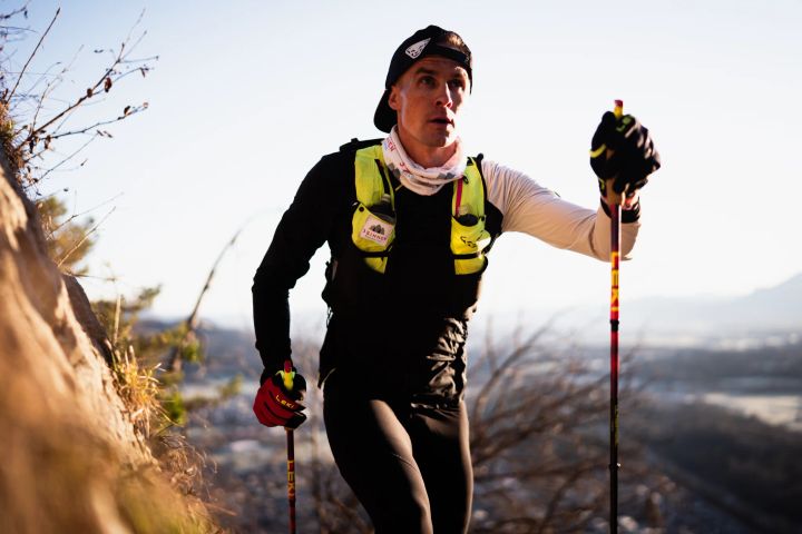 Trailrunner in langer schwarzer Tight, Laufweste und Kappe steigt mit Stöcken einen Hang hinauf, dahinter ein Tal im Gegenlicht
