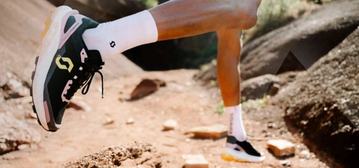 Läufer in weißen Socken und dunklem Trailschuh mit gelbem Logo springt über einen staubigen Pfad zwischen Felsen