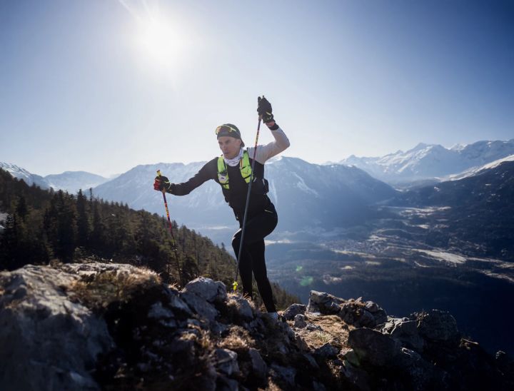 Trailläufer mit Stöcken und gelber Laufweste auf einem Felsgrat in der Sonne, dahinter ein Tal und verschneite Berge