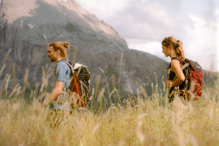 Wanderer in hellblauem T-Shirt und Wanderin in dunklem Top gehen mit Rucksäcken durch eine Wiese mit hohen Gräsern vor Felswänden
