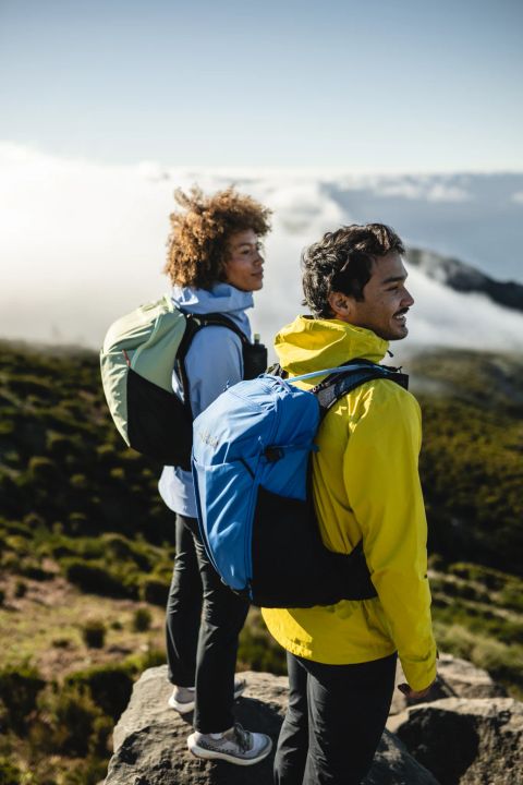 Zwei Wandernde mit Tagesrucksäcken stehen auf einem Felsen über den Wolken
