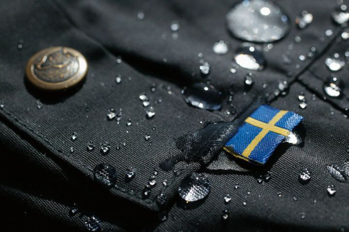Nahaufnahme von dunklem G-1000-Stoff mit Wassertropfen, Druckknopf und kleiner schwedischer Flagge