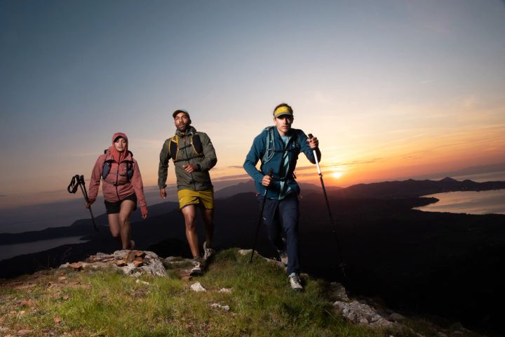 Drei Wandernde mit Stöcken auf einem Grat im Abendlicht, der Mann in der Mitte trägt eine gelbe Shorts
