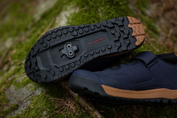 Radschuh mit Gummiprofil von unten auf einem bemoosten Stein, in der Mitte ein versenkt montiertes 2-Loch-Cleat, daneben ein zweiter dunkelblauer Schuh