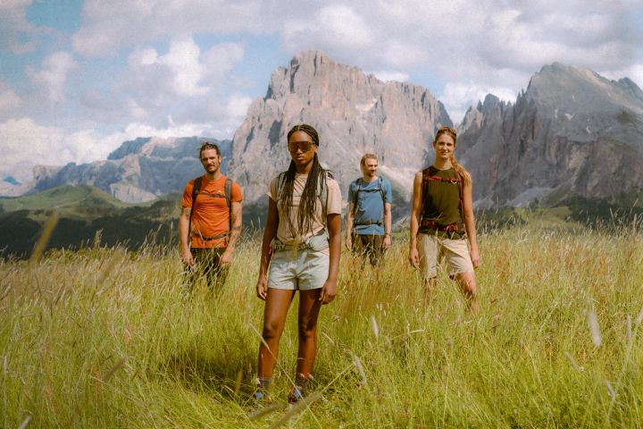 Vier Wandernde in T-Shirts, Tops und Shorts mit Rucksäcken stehen in einer Wiese mit hohen Gräsern, dahinter schroffe Felsgipfel