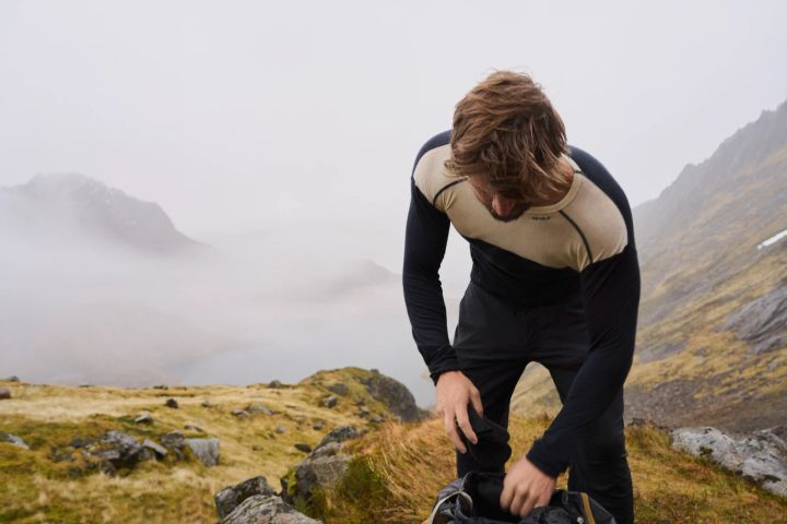 Person in schwarz-beigem Langarmshirt und schwarzer Hose beugt sich auf einer Bergwiese über ihren Rucksack, dahinter Nebel