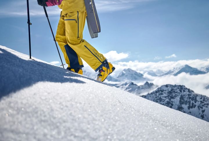 Person in gelber Hose und gelben Skitourenschuhen steigt mit Stöcken einen Schneehang hinauf, am Rucksack ein Ski, dahinter Gipfel über den Wolken