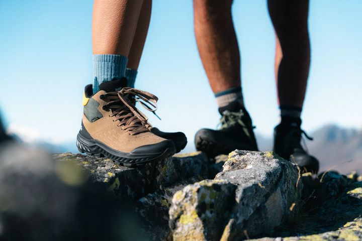 Zwei Wanderer stehen auf einem Felsgrat, vorn ein brauner knöchelhoher Wanderschuh mit hellblauer Socke, dahinter schwarze Schuhe und blauer Himmel