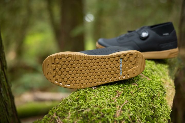 Schwarzer Flatpedal-Schuh mit honigfarbener Gummisohle mit Wabenprofil liegt auf einem bemoosten Baumstamm, dahinter ein zweiter Schuh
